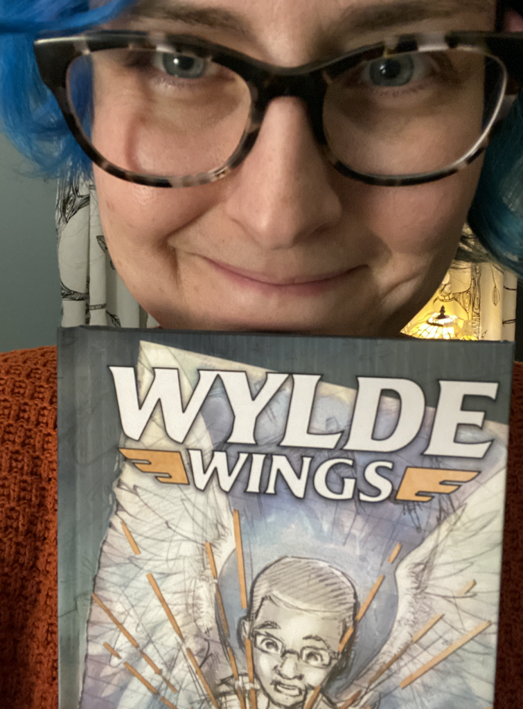 Happy Kate lady with blue hair and big glasses and a smile with Wylde Wings book
